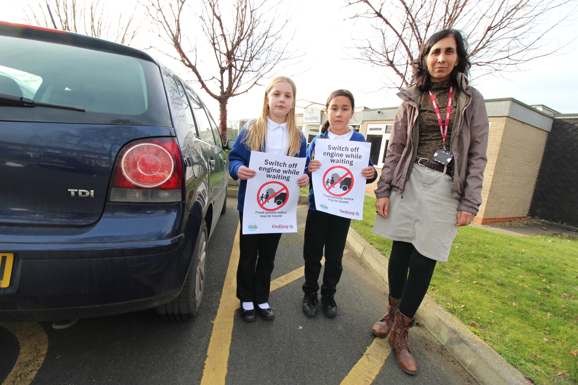 Left to Right – Faye Garrard and Lyra Jagessar from Ernhale Junior School with Gedling Borough Council Climate Change Officer Sim Duhra stood in a school car park next to a dark blue car. Both School children are holding signs which read 